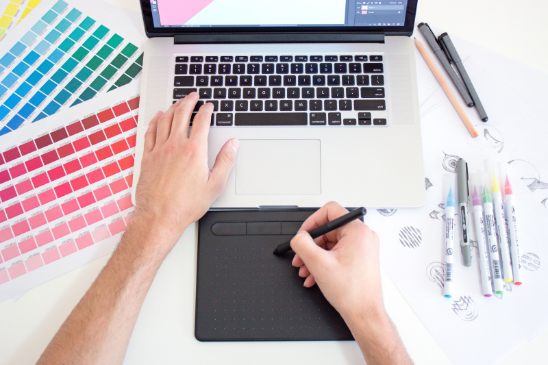   Grafisk designer som arbetar på en Macbook-dator med hjälp av en styrplatta, färgdiagram och markörer